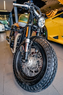 A close-up view of a custom motorcycle's front tire and headlight. The bike has a vintage design, featuring a matte green gas tank with a yellow stripe, and a prominent, thick black tire with detailed spokes. In the background, there is a bright yellow sports car with sleek, modern lines, parked in a garage or showroom setting.