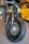 A close-up view of a custom motorcycle's front tire and headlight. The bike has a vintage design, featuring a matte green gas tank with a yellow stripe, and a prominent, thick black tire with detailed spokes. In the background, there is a bright yellow sports car with sleek, modern lines, parked in a garage or showroom setting.