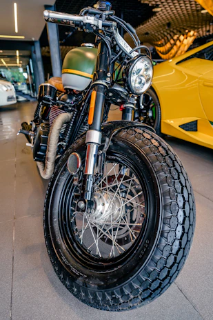 A close-up view of a custom motorcycle's front tire and headlight. The bike has a vintage design, featuring a matte green gas tank with a yellow stripe, and a prominent, thick black tire with detailed spokes. In the background, there is a bright yellow sports car with sleek, modern lines, parked in a garage or showroom setting.