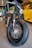 A close-up view of a custom motorcycle's front tire and headlight. The bike has a vintage design, featuring a matte green gas tank with a yellow stripe, and a prominent, thick black tire with detailed spokes. In the background, there is a bright yellow sports car with sleek, modern lines, parked in a garage or showroom setting.