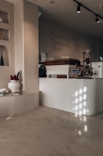 A cozy café interior with minimalist design. The counter, labeled 'ARTEA CAFE,' features a sleek, modern look with a light wooden finish. Above, there are overhead track lights. A variety of coffee options is listed on the wall, including Flat White, Handbrew, and Shot. On the side, there is a decorative shelf with vases, flowers in a white pot, and some books. The play of light creates an inviting atmosphere with soft shadows on the smooth, neutral-toned walls and floors.