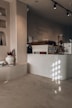 A cozy caf&eacute; interior with minimalist design. The counter, labeled 'ARTEA CAFE,' features a sleek, modern look with a light wooden finish. Above, there are overhead track lights. A variety of coffee options is listed on the wall, including Flat White, Handbrew, and Shot. On the side, there is a decorative shelf with vases, flowers in a white pot, and some books. The play of light creates an inviting atmosphere with soft shadows on the smooth, neutral-toned walls and floors.