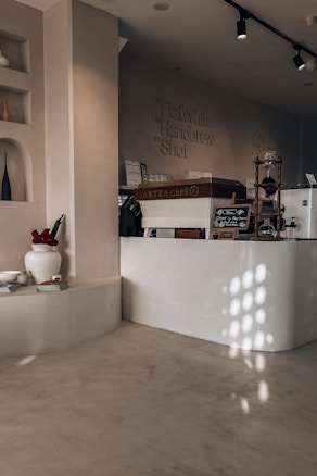 A cozy caf&eacute; interior with minimalist design. The counter, labeled 'ARTEA CAFE,' features a sleek, modern look with a light wooden finish. Above, there are overhead track lights. A variety of coffee options is listed on the wall, including Flat White, Handbrew, and Shot. On the side, there is a decorative shelf with vases, flowers in a white pot, and some books. The play of light creates an inviting atmosphere with soft shadows on the smooth, neutral-toned walls and floors.