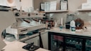 A well-equipped coffee bar with a professional espresso machine, cups, milk frother, and various bottles and containers. Shelves display certificates and a blender, and a mini-fridge is stocked with beverages. There's a milk carton and tools for tamping coffee nearby.