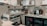 A well-equipped coffee bar with a professional espresso machine, cups, milk frother, and various bottles and containers. Shelves display certificates and a blender, and a mini-fridge is stocked with beverages. There's a milk carton and tools for tamping coffee nearby.