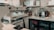 A well-equipped coffee bar with a professional espresso machine, cups, milk frother, and various bottles and containers. Shelves display certificates and a blender, and a mini-fridge is stocked with beverages. There's a milk carton and tools for tamping coffee nearby.