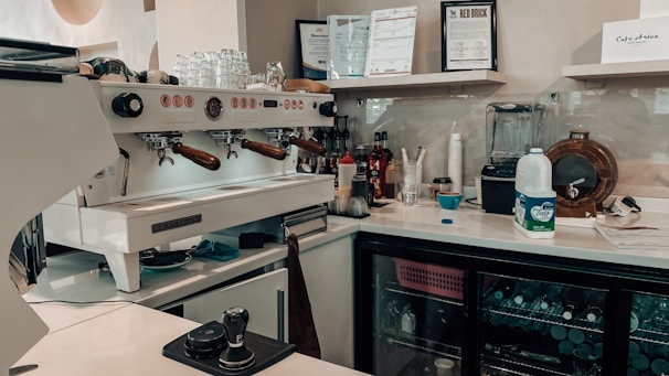 A well-equipped coffee bar with a professional espresso machine, cups, milk frother, and various bottles and containers. Shelves display certificates and a blender, and a mini-fridge is stocked with beverages. There's a milk carton and tools for tamping coffee nearby.