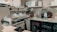 A well-equipped coffee bar with a professional espresso machine, cups, milk frother, and various bottles and containers. Shelves display certificates and a blender, and a mini-fridge is stocked with beverages. There's a milk carton and tools for tamping coffee nearby.
