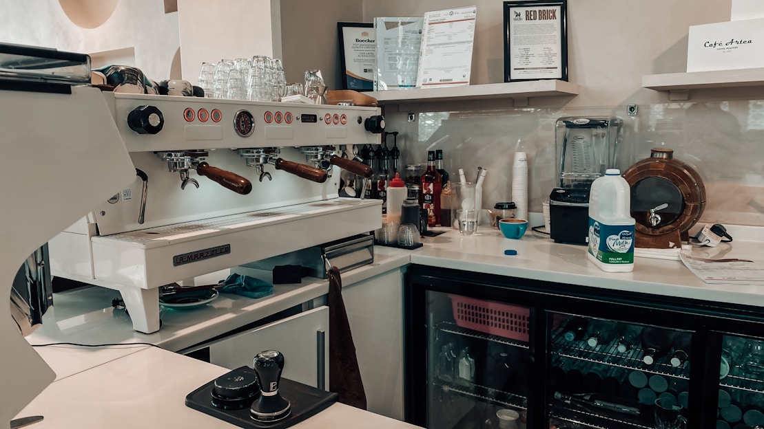A well-equipped coffee bar with a professional espresso machine, cups, milk frother, and various bottles and containers. Shelves display certificates and a blender, and a mini-fridge is stocked with beverages. There's a milk carton and tools for tamping coffee nearby.
