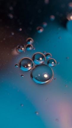 Clusters of transparent oil bubbles float on a smooth, blue surface, casting reflections and slight shadows. The larger bubbles are surrounded by smaller ones, displaying a clear spherical shape with reflective highlights.