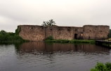 A rustic stone fortress stands by the edge of a reflective body of water, surrounded by lush greenery. The fortress features weathered stone walls with small openings that resemble windows. A wooden bridge extends over the water, leading to the fortress entrance. The sky is overcast, suggesting a calm, tranquil atmosphere.