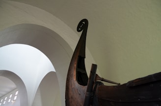 The image captures the detailed upper structure of an intricately carved wooden boat with a large, spiraled bow. The surroundings include a spacious, arched, light-colored interior, suggesting a museum setting.