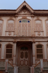 A symmetrical, historic building with ornate architectural details and a vintage aesthetic. It features tall columns, large decorative windows, and an intricate facade. The wooden door is flanked by stairs leading up to it, and there are signs of wear and age on the structure.