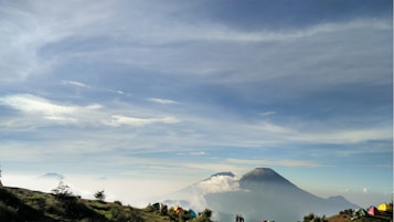 A serene landscape features a large mountain with its peak shrouded in clouds, set against a backdrop of expansive blue sky with wispy clouds. In the foreground, there are clusters of colorful tents and a few small groups of people possibly engaged in hiking or camping.