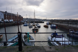 A harbor with several small boats and yachts docked on calm water. The scene includes a stone-paved pier with a metal railing in the foreground, where a person is seen stepping onto a boat. On the left, there is a row of brick buildings with boats stored alongside. The background shows a cloudy sky with muted lighting, casting reflections on the water.