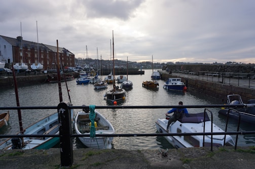 A harbor with several small boats and yachts docked on calm water. The scene includes a stone-paved pier with a metal railing in the foreground, where a person is seen stepping onto a boat. On the left, there is a row of brick buildings with boats stored alongside. The background shows a cloudy sky with muted lighting, casting reflections on the water.