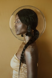 A woman with dark skin and long braided hair is seen in profile. She wears large tassel earrings and a shimmering gold garment that matches the warm, monochromatic background. A circular halo-like object frames her head, and her skin glistens with a subtle, metallic sheen.