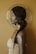 A woman with dark skin and long braided hair is seen in profile. She wears large tassel earrings and a shimmering gold garment that matches the warm, monochromatic background. A circular halo-like object frames her head, and her skin glistens with a subtle, metallic sheen.