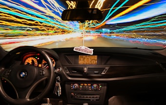 A car interior is seen with a focus on the steering wheel and dashboard. Outside the windshield, vibrant streaks of light create a dynamic, colorful motion effect, suggesting high speed or long exposure photography. The dashboard displays illuminated gauges, and a car-shaped air freshener hangs from the rearview mirror.