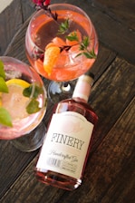 An arrangement of three gin cocktails in large glasses, garnished with fruit slices and herbs, sits on a wooden table. A bottle labeled 'Finery Handcrafted Gin' is placed alongside the glasses, indicating the brand of the gin used.