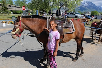 A young girl stands beside a saddled brown horse on a sunny day. They are in an outdoor setting with a stop sign, trees, and mountains visible in the background. Several people and vehicles are in the distance, indicating a lively area.