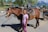 A young girl happily riding a horse in a sunny outdoor arena.