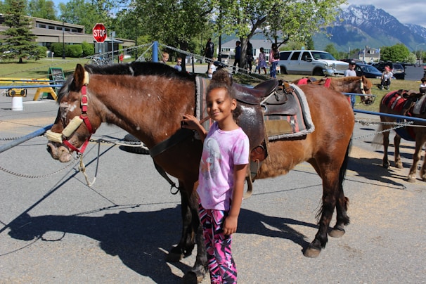 A young girl stands beside a saddled brown horse on a sunny day. They are in an outdoor setting with a stop sign, trees, and mountains visible in the background. Several people and vehicles are in the distance, indicating a lively area.
