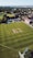 Aerial view of a cricket field with players wearing uniforms, engaging in a match. The field is surrounded by green grass, with a few buildings and trees visible in the background under a clear sky.