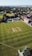 Aerial view of a cricket field with players wearing uniforms, engaging in a match. The field is surrounded by green grass, with a few buildings and trees visible in the background under a clear sky.