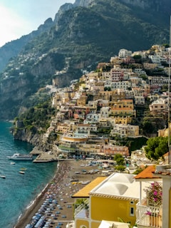 A picturesque coastal town with colorful buildings built into the cliffs, overlooking a vibrant beach lined with umbrellas and people. The blue sea contrasts with the rugged mountains in the background, creating a harmonious natural setting.