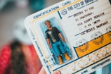 A toy figure of a man with distinctive hairstyles in a plastic packaging is placed on top of a card that resembles an identification document. The card includes stylized text and graphics, mimicking an official-looking document from 'The City of Brooklyn'. It contains playful artistic elements, vibrant colors, and creative typography.