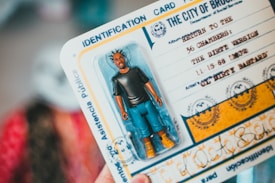 A toy figure of a man with distinctive hairstyles in a plastic packaging is placed on top of a card that resembles an identification document. The card includes stylized text and graphics, mimicking an official-looking document from 'The City of Brooklyn'. It contains playful artistic elements, vibrant colors, and creative typography.