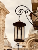 A vintage street lamp hangs from an ornate black metal bracket in front of an architectural backdrop featuring classical buildings with domes and intricate details.