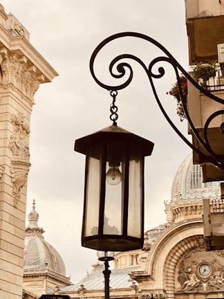 A vintage street lamp hangs from an ornate black metal bracket in front of an architectural backdrop featuring classical buildings with domes and intricate details.