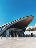 A modern transportation hub exterior with an arched roof structure dominates the scene. People are entering and exiting through glass doors beneath the vast curve. The architecture features sleek, contemporary lines with a combination of glass and metal materials.