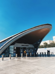 A modern transportation hub exterior with an arched roof structure dominates the scene. People are entering and exiting through glass doors beneath the vast curve. The architecture features sleek, contemporary lines with a combination of glass and metal materials.