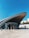 A modern transportation hub exterior with an arched roof structure dominates the scene. People are entering and exiting through glass doors beneath the vast curve. The architecture features sleek, contemporary lines with a combination of glass and metal materials.