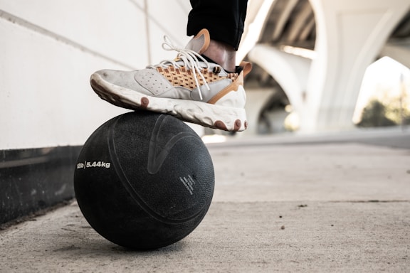 A person balances one foot atop a large black medicine ball weighing 5.44kg. The person is wearing stylish, multi-colored sneakers with white, gray, and brown accents. The scene is set outdoors beneath an overpass, with concrete pavement and the underside of the structure visible.