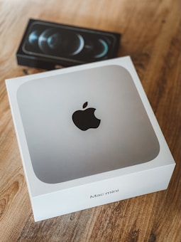 A Mac Mini box with a sleek Apple logo on top is placed on a wooden table. Behind it, there is a partially obscured box for a smartphone, possibly an iPhone, featuring a design of three camera lenses on the packaging.