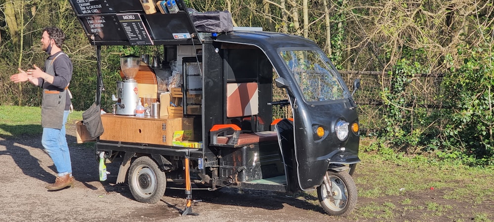 A man wearing blue jeans, brown shoes, and a grey apron is standing next to a three-wheeled vehicle that has been converted into a mobile coffee cart. The vehicle has various coffee-making equipment and supplies on it, including a grinder, cups, and a menu board attached to the roof. The background features grassy and wooded areas with a fence.