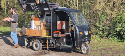 A man wearing blue jeans, brown shoes, and a grey apron is standing next to a three-wheeled vehicle that has been converted into a mobile coffee cart. The vehicle has various coffee-making equipment and supplies on it, including a grinder, cups, and a menu board attached to the roof. The background features grassy and wooded areas with a fence.