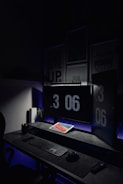 A dark, minimalist workspace is illuminated by two large computer monitors displaying the time as 3:06 PM. A tablet lies on the desk, displaying a colorful screen. The setup includes a keyboard and mouse on a smooth surface. Subdued lighting and motivational posters create a focused atmosphere.