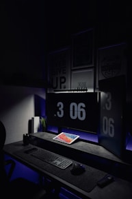 A dark, minimalist workspace is illuminated by two large computer monitors displaying the time as 3:06 PM. A tablet lies on the desk, displaying a colorful screen. The setup includes a keyboard and mouse on a smooth surface. Subdued lighting and motivational posters create a focused atmosphere.