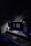 A dark, minimalist workspace is illuminated by two large computer monitors displaying the time as 3:06 PM. A tablet lies on the desk, displaying a colorful screen. The setup includes a keyboard and mouse on a smooth surface. Subdued lighting and motivational posters create a focused atmosphere.