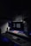A dark, minimalist workspace is illuminated by two large computer monitors displaying the time as 3:06 PM. A tablet lies on the desk, displaying a colorful screen. The setup includes a keyboard and mouse on a smooth surface. Subdued lighting and motivational posters create a focused atmosphere.