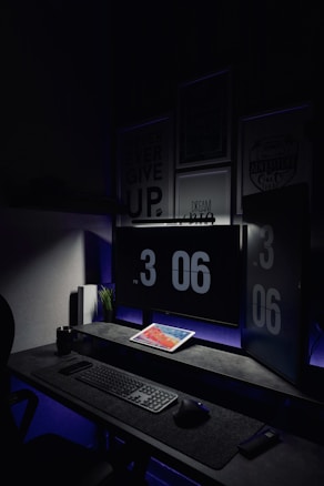 A dark, minimalist workspace is illuminated by two large computer monitors displaying the time as 3:06 PM. A tablet lies on the desk, displaying a colorful screen. The setup includes a keyboard and mouse on a smooth surface. Subdued lighting and motivational posters create a focused atmosphere.