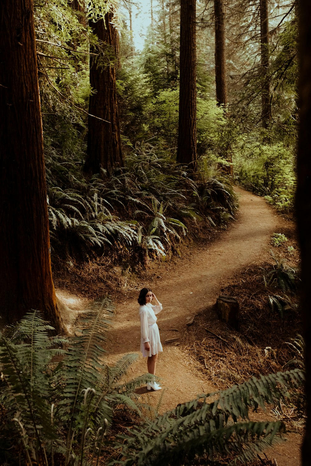 girl in white shirt walking on dirt road between green plants and trees during daytime