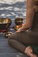 A person sits cross-legged on a wooden floor, surrounded by several singing bowls and a mallet. The person is dressed in brown leggings and a sleeveless top. The scene is bathed in dappled natural light, creating a serene and meditative atmosphere.