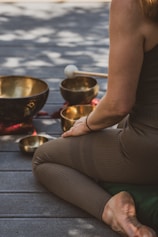 A person sits cross-legged on a wooden floor, surrounded by several singing bowls and a mallet. The person is dressed in brown leggings and a sleeveless top. The scene is bathed in dappled natural light, creating a serene and meditative atmosphere.