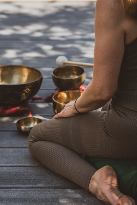 A person sits cross-legged on a wooden floor, surrounded by several singing bowls and a mallet. The person is dressed in brown leggings and a sleeveless top. The scene is bathed in dappled natural light, creating a serene and meditative atmosphere.